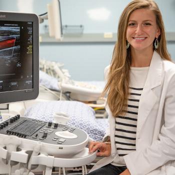 Female student wearing a lab coat sitting next to an ultrasound machine.